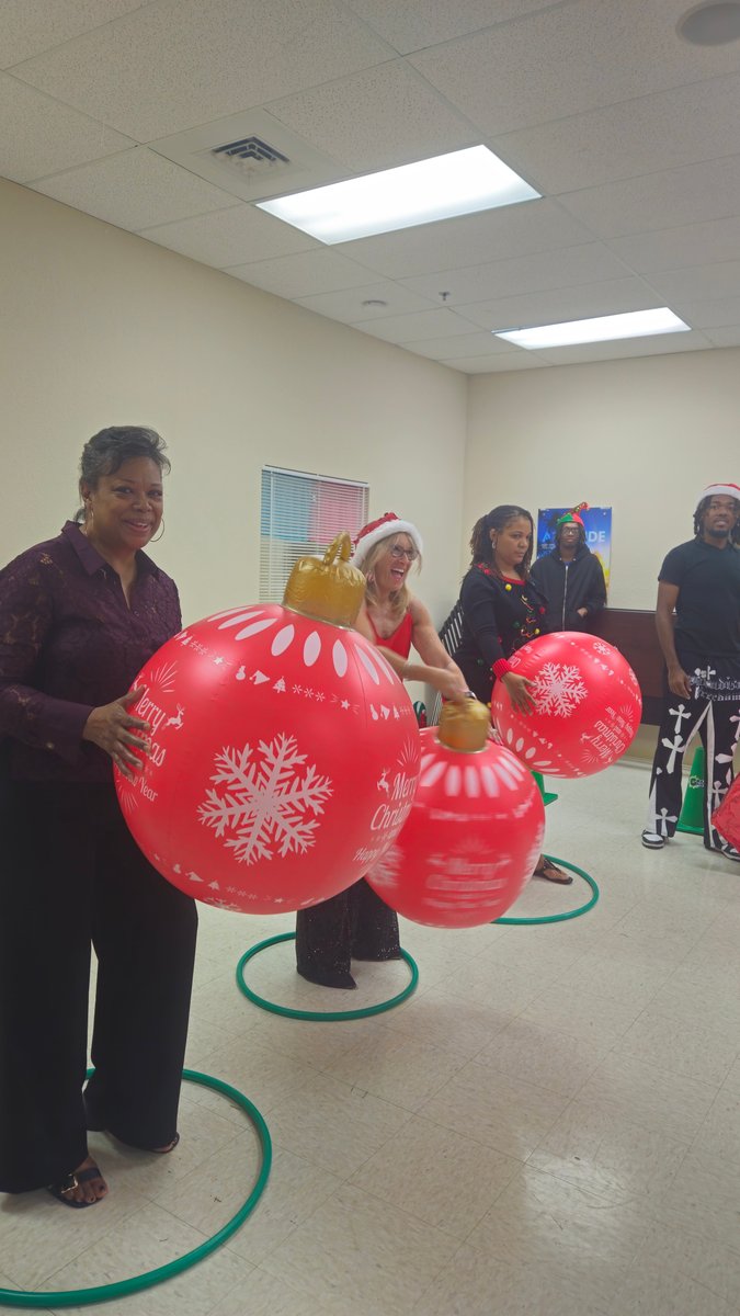 Adults carrying giant red Christmas ornaments in a relay race inside a hall during a holiday family night