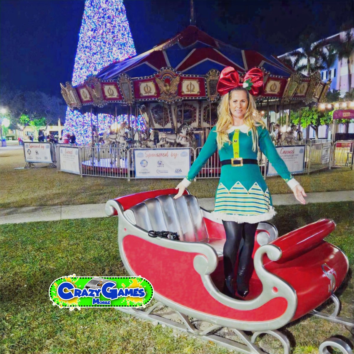 Crazy Games Christmas Elf character standing in a red sleigh prop at a night holiday fair with a lit tree behind