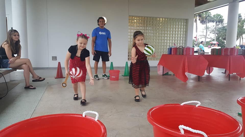Kids running the Crazy Games FL Giant Ornament and Spoon Race with red buckets under a covered pavilion at a holiday party in Palm Beach County