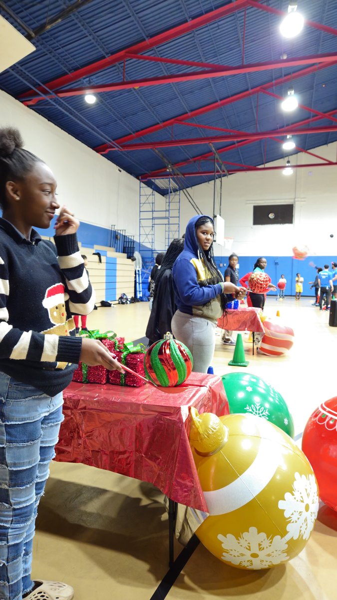 A student picking up a giant ornament from a holiday games table set with gifts and red tablecloths in a school gym in West Palm Beach