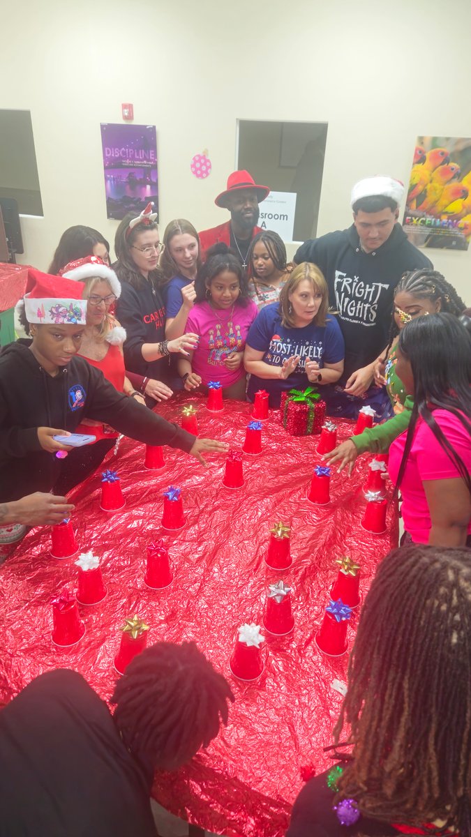 Kids, teens and parents in Christmas hats crowded around a long red table playing a holiday cup game at an indoor holiday party