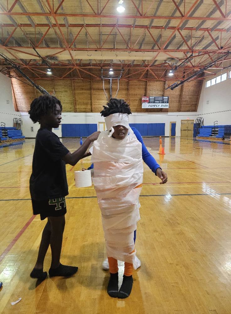 Kids playing a mummy wrap race on a gym floor at an indoor Halloween-style party