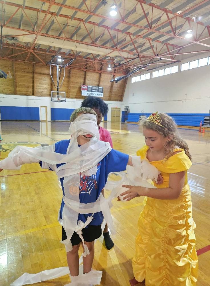 Kids in costumes doing a mummy wrap relay at a Halloween party in a school gym in South Florida