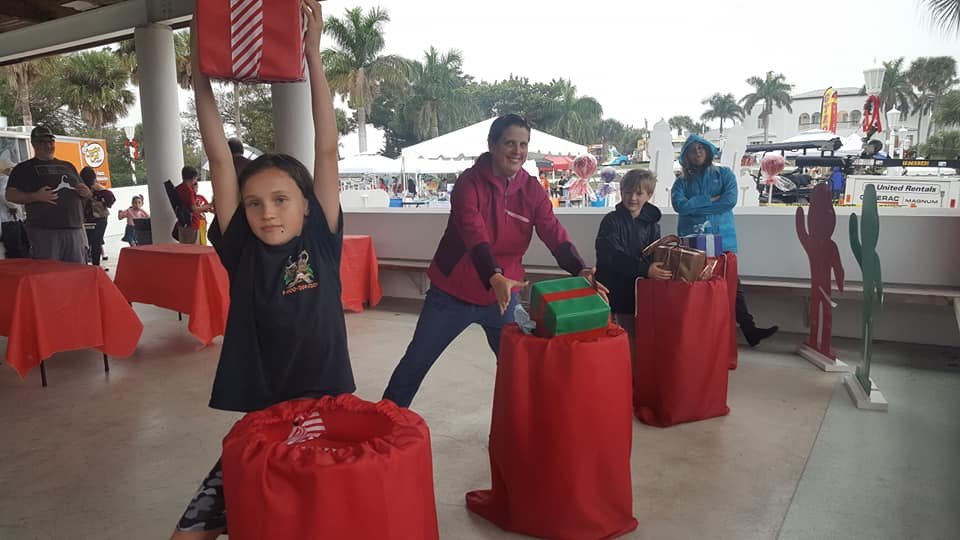 Kids playing the Crazy Games FL Santa's Helper gift relay game under an outdoor pavilion with palm trees