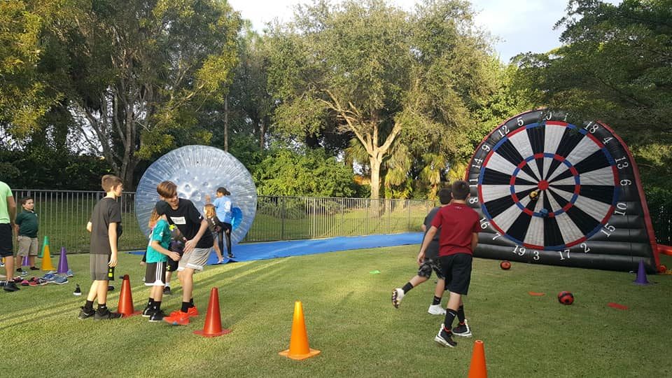 Kids kicking balls at the giant Soccer Target with a Human Zorb Ball nearby at a park event in Palm Beach County