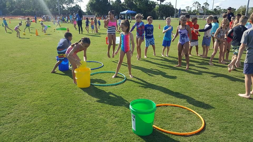 Kids running a sponge relay with buckets and hula hoops on a grass field at a South Florida party